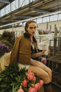 model im brauner workwearjacke sitz auf Metallgestell und hält einen Strauss lila Blumen im Blumenladen