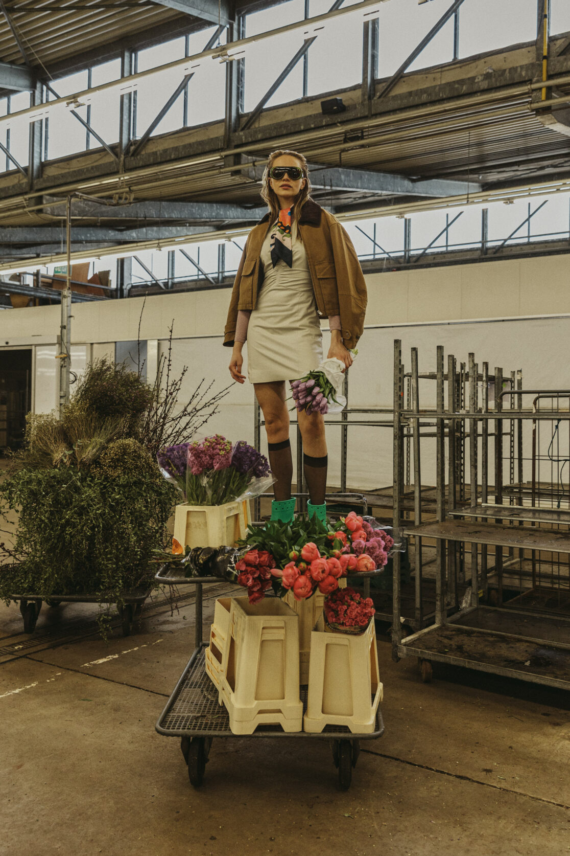 Model im beigen Trenchcoat und Overknee-Socken steht zwischen Metallgestellen in einer Gärtnerei