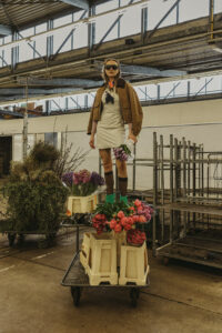 Model im beigen Trenchcoat und Overknee-Socken steht zwischen Metallgestellen in einer Gärtnerei