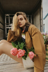 model sitzt mit brauner workwearjacke auf einer LKW Ladefläche mit einem Strauss pinker pfingsrosen