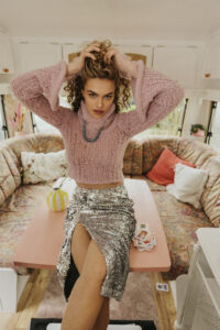 Model sitzt im Glitzer Rock und rosa Strick Pullover im Innenraum des Wohnwagens auf dem Esstisch und hält sich die Haare hoch