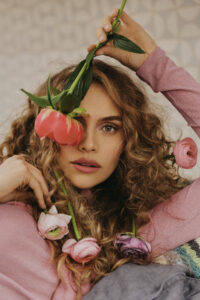 Model im rosa Oberteil hat bunte Blumen im Haar verteilt und hält eine Blume vor ihrem Augen